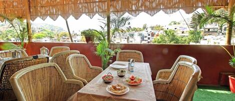 An outdoor dining area with a table set for two, surrounded by wicker chairs, under a thatched roof.