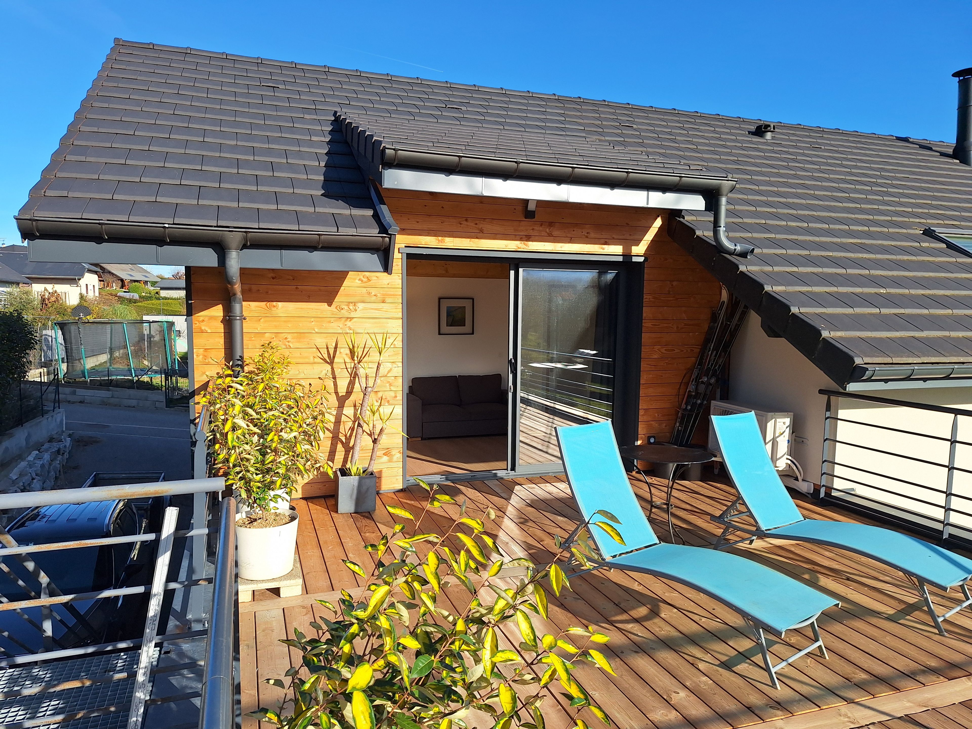 Een modern huis met een houten terras, een schuifdeur die naar het interieur leidt, en een blauwe buitenstoel.