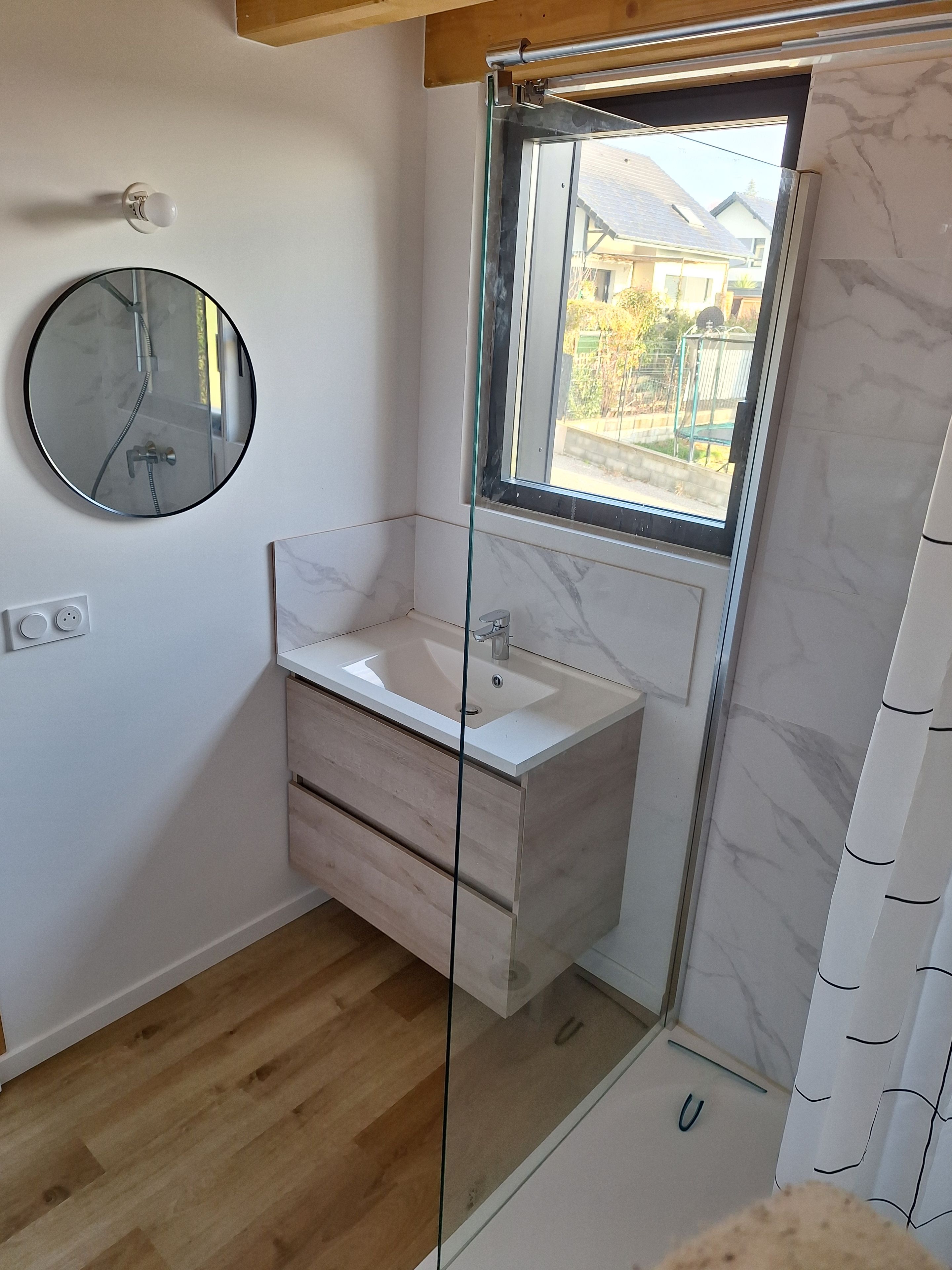 Une salle de bain avec une douche à parois de verre, un miroir rond et un lavabo blanc doté de tiroirs en bois.