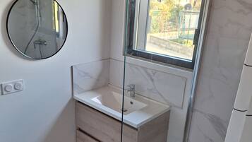 Une salle de bain avec une douche à parois de verre, un miroir rond et un lavabo blanc doté de tiroirs en bois.