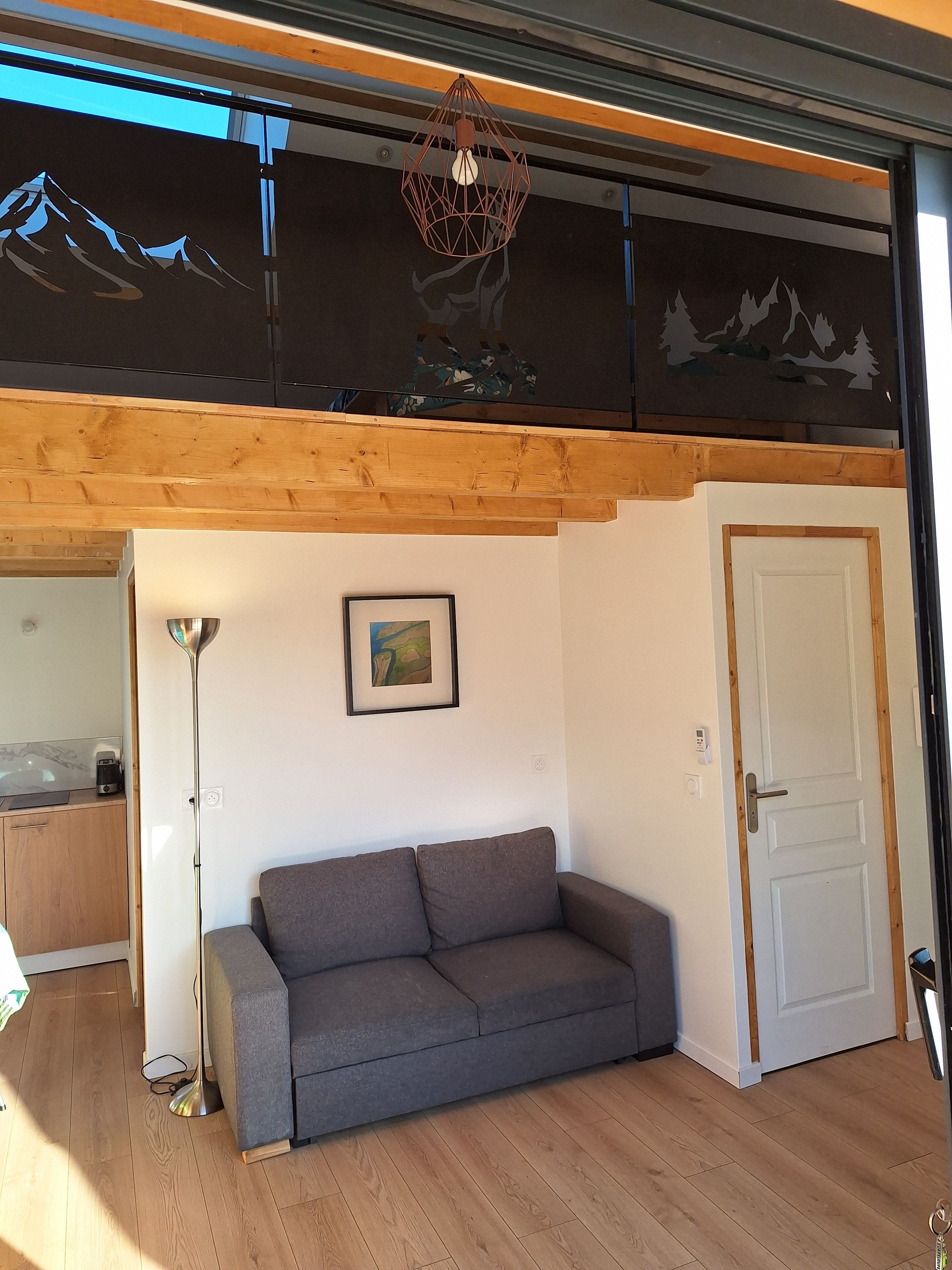 Een kamer met een grijze bank, een lijst met een foto aan de muur, een deur en een plafondlamp.