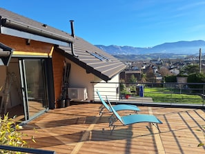 A residential property with a wooden deck, a glass door entrance, a chimney, and a view of a town and mountains in the background. - La Montagnette - With a terrace overlooking the mountains (Entrelacs)