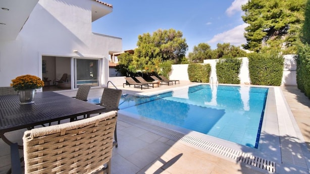 A modern outdoor pool area with a dining table and chairs, surrounded by greenery and a clear sky.