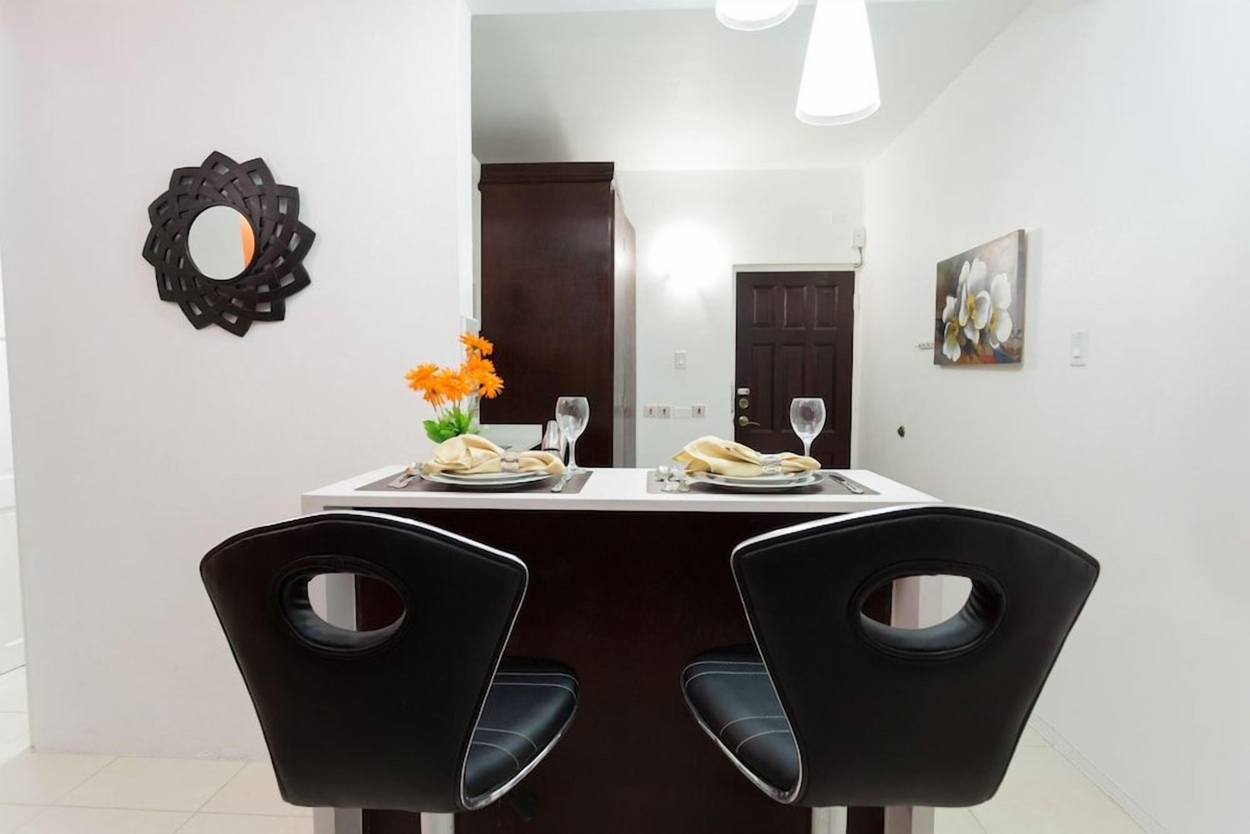 A dining area with a table set for two, two black chairs with cut-out backrests, a mirror, and a painting on the wall.