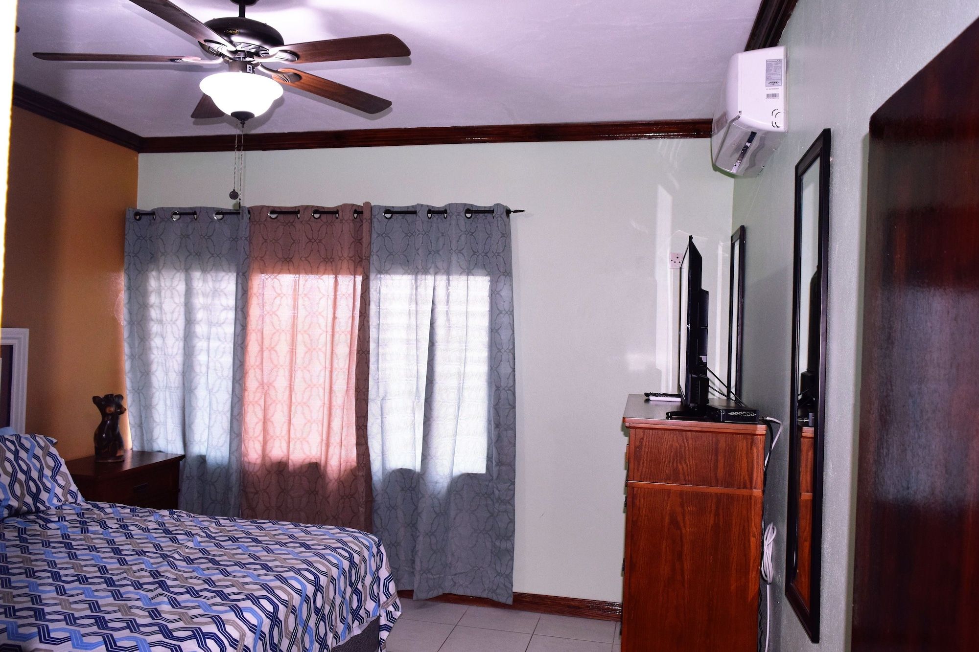 Une chambre à coucher avec un lit, un ventilateur de plafond, un téléviseur posé sur un meuble en bois et une fenêtre avec des rideaux.