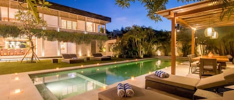 Un área junto a la piscina con sillones de descanso, una mesa de comedor y una pérgola con luces.