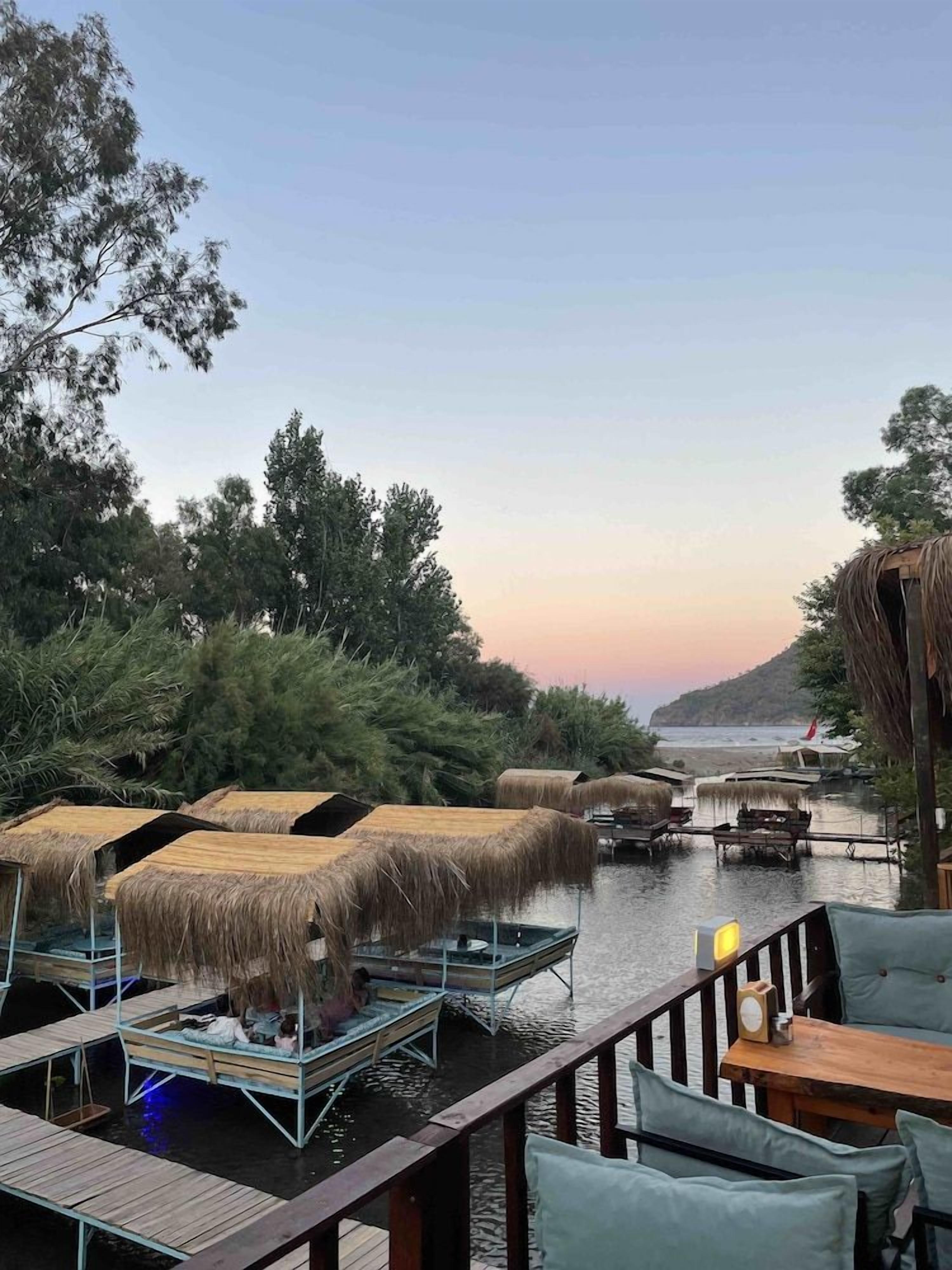 Eine Uferterrasse mit Reetdachschirmen, Holzmöbeln und Blick auf das Wasser und das entfernte Land.