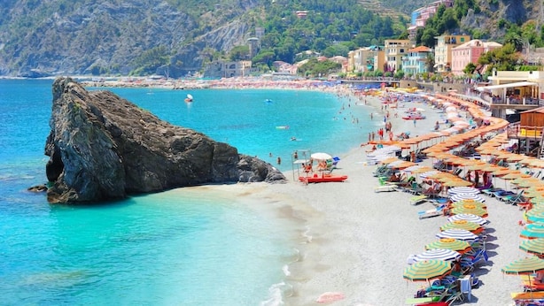 A beach with a rocky outcrop, a row of colorful umbrellas, and buildings in the background. - The First Riomaggiore 5 Terre (Riomaggiore)