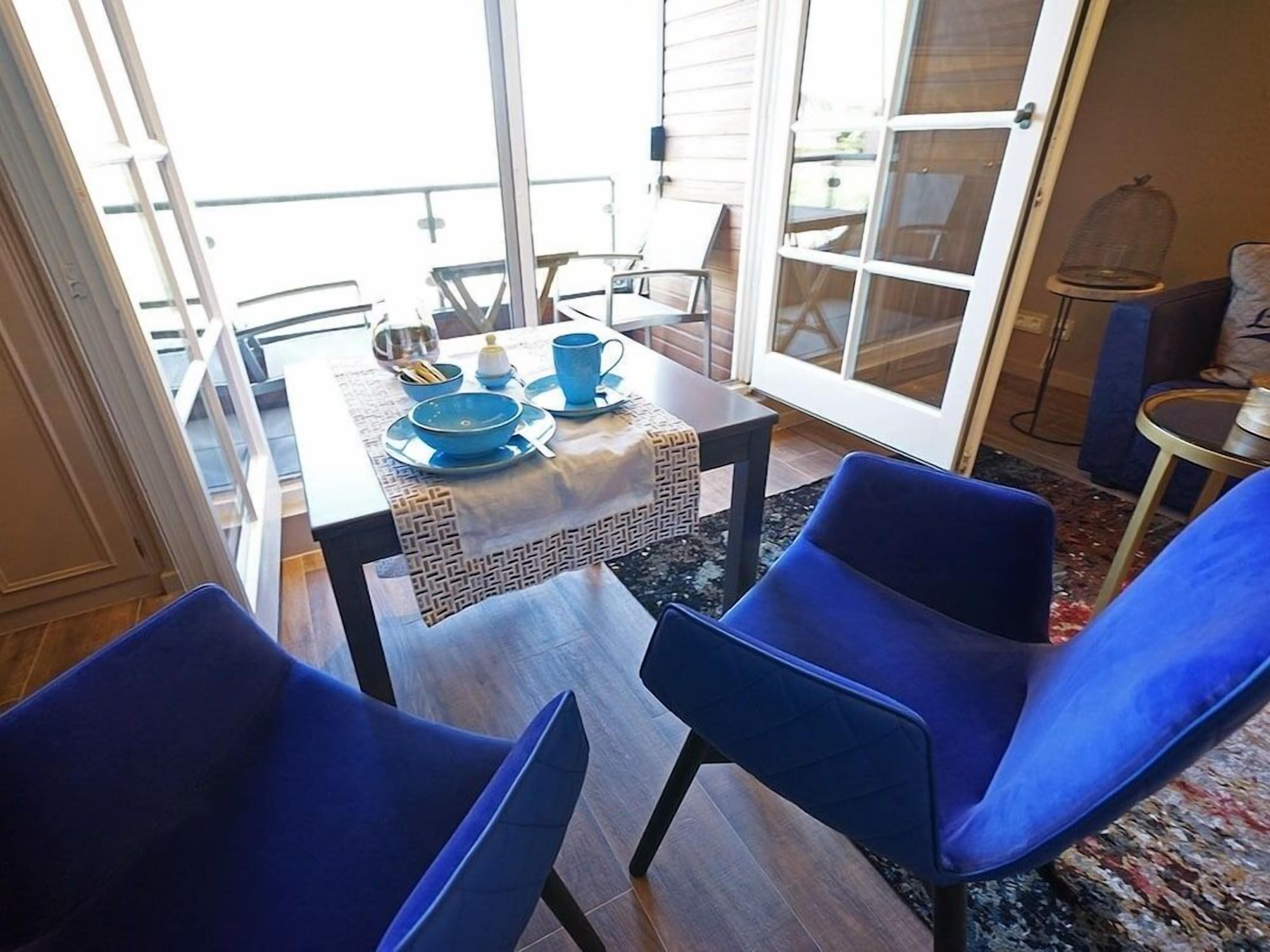 Zona pranzo con tavolo apparecchiato per due, sedie blu e vista attraverso una porta vetrata.