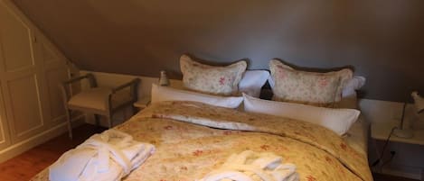 A neatly made bed with floral bedding and two sets of towels, a chair, and a bedside table with a lamp.