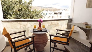 Un balcón con dos sillas, una mesa con alimentos para el desayuno y vistas a edificios y árboles.