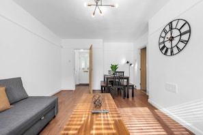 Una sala de estar moderna con un sofá, una mesa de comedor con sillas, un reloj de pared grande y una escalera que lleva a otra habitación.