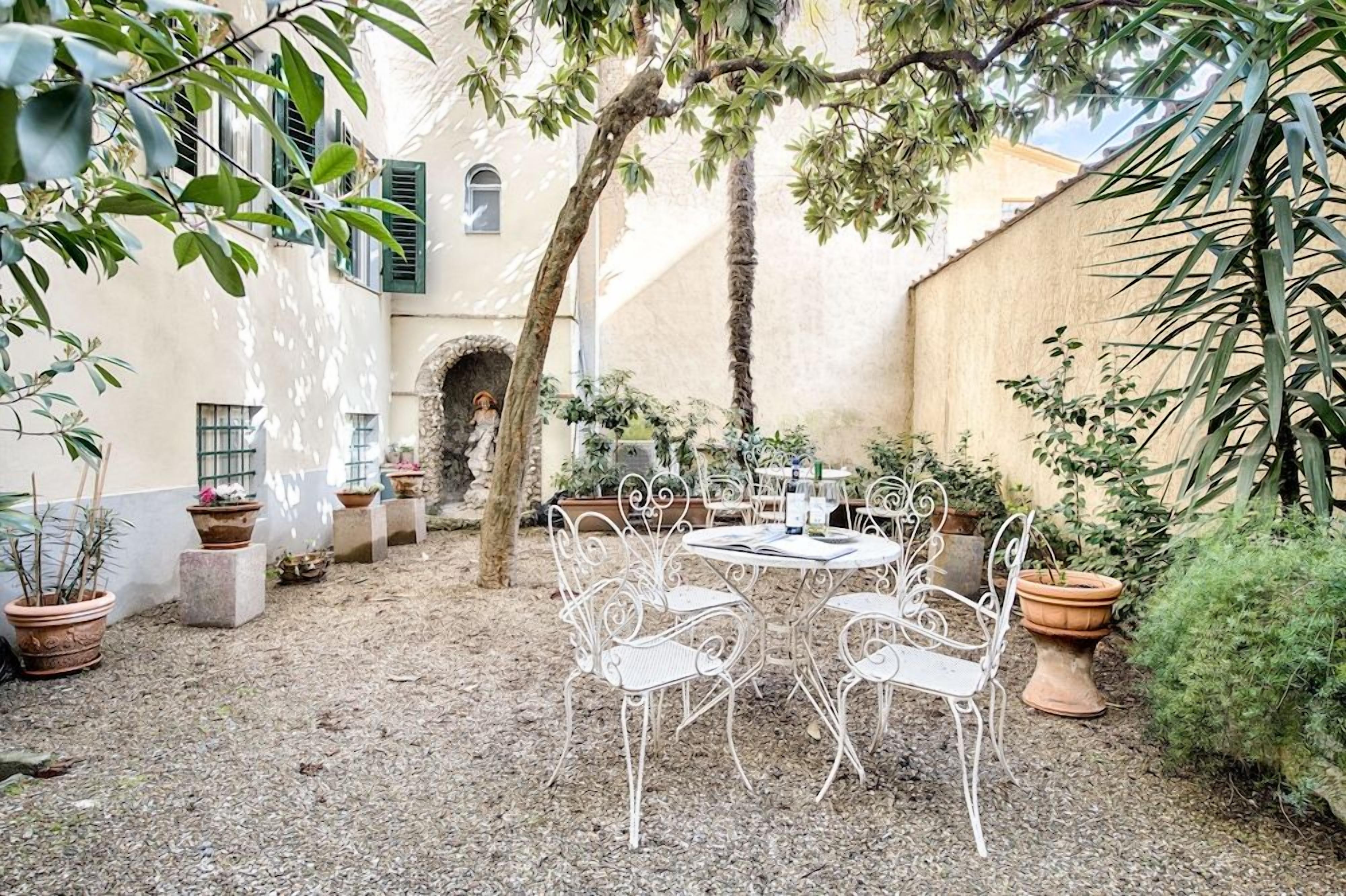 An outdoor patio with white metal chairs and a table, surrounded by potted plants and a tree.