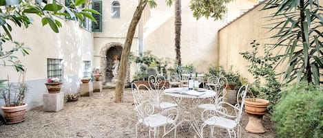 Un patio al aire libre con sillas y una mesa de metal blanco, rodeado de plantas en macetas y un árbol.