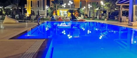 Une piscine dont le bord est illuminé la nuit, entourée d’un bâtiment doté de nombreux balcons et d’espaces de détente en plein air.