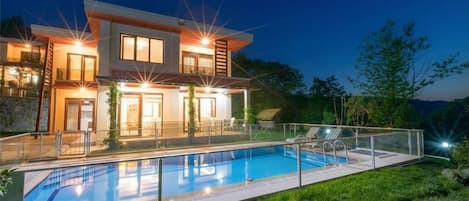 Une maison moderne à deux étages, dotée d’une piscine, de mobilier d’extérieur et d’un éclairage extérieur soigné.