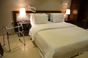 A hotel room with a bed, two bedside lamps, a nightstand, a wall-mounted handrail, and a walking aid.