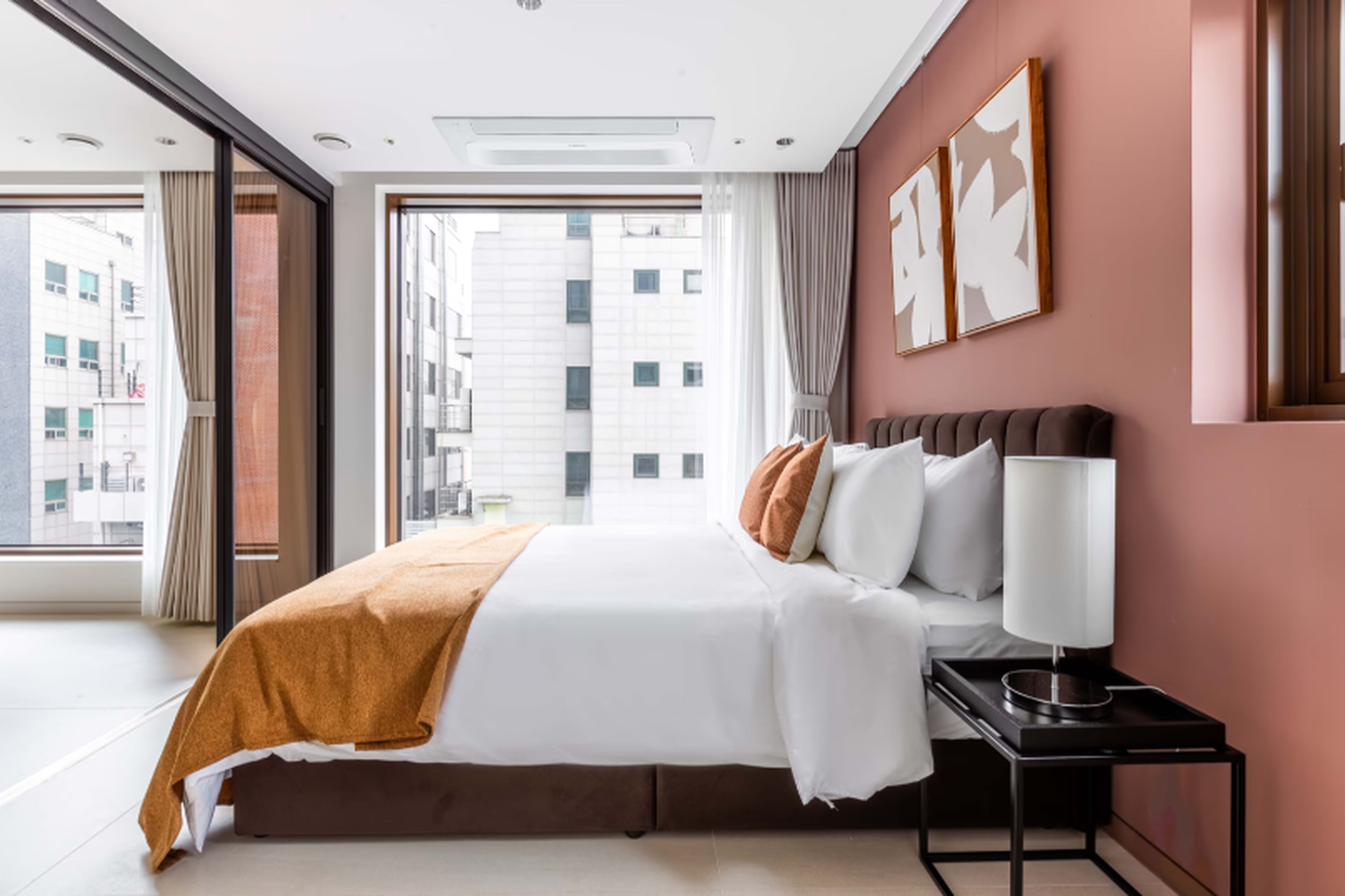 A hotel room with a large bed, a bedside table with a lamp, and a view of the city through a large window.