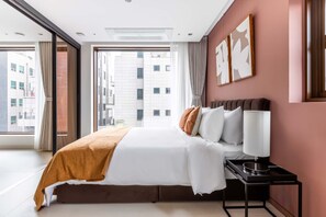 A hotel room with a large bed, a bedside table with a lamp, and a view of the city through a large window. - Luxia Dosan - Executive Suite - Fully Furnished 1BR 1B (Seoul)