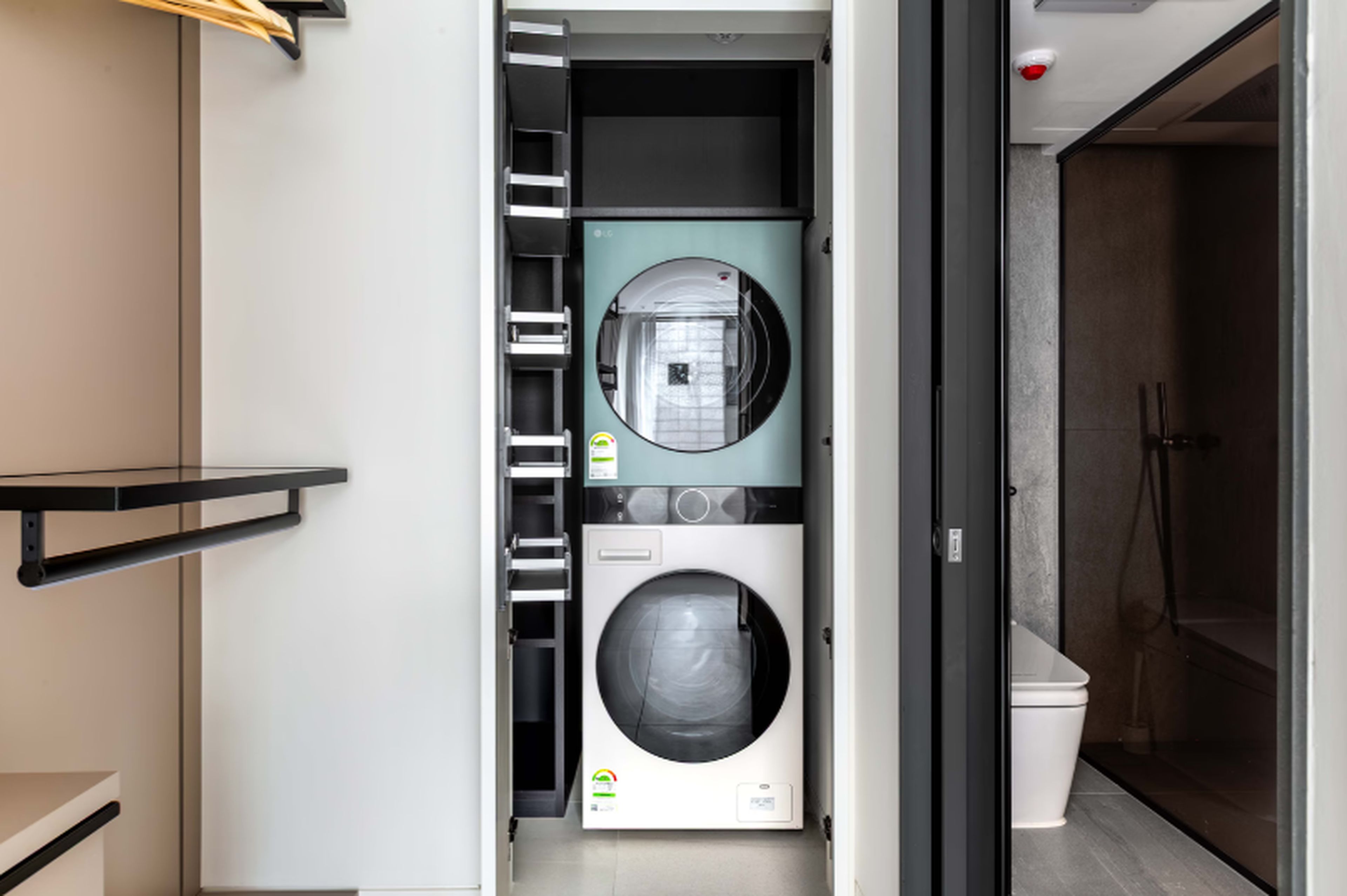 A compact laundry room with a front-loading washer and dryer, a small shelf, and a door leading to a shower area.