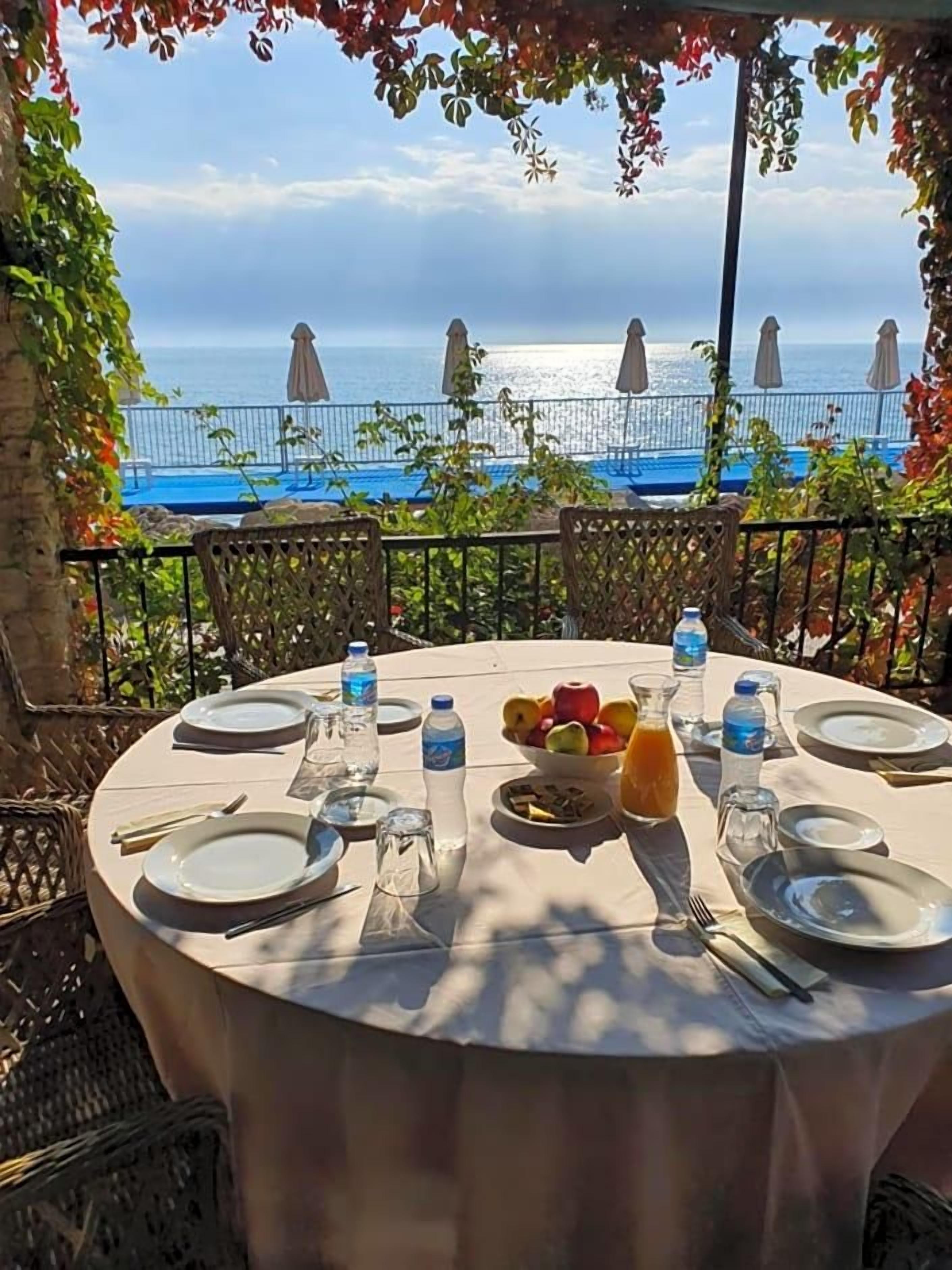 A round table set for a meal with a view of the sea, surrounded by chairs and umbrellas.