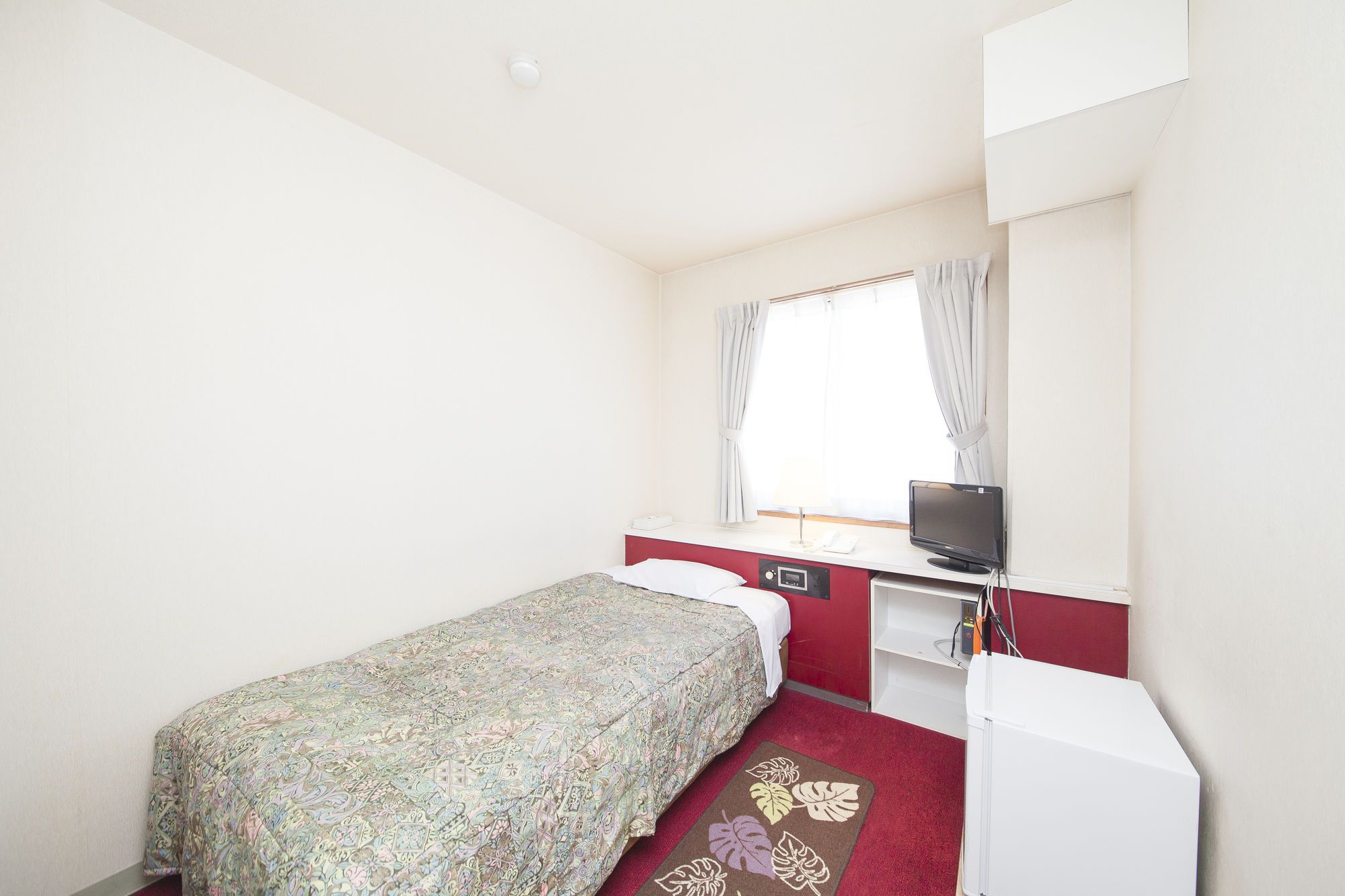 A small, single-bed room with a red and white color scheme, a desk with a computer, a window with curtains, and a small white refrigerator.