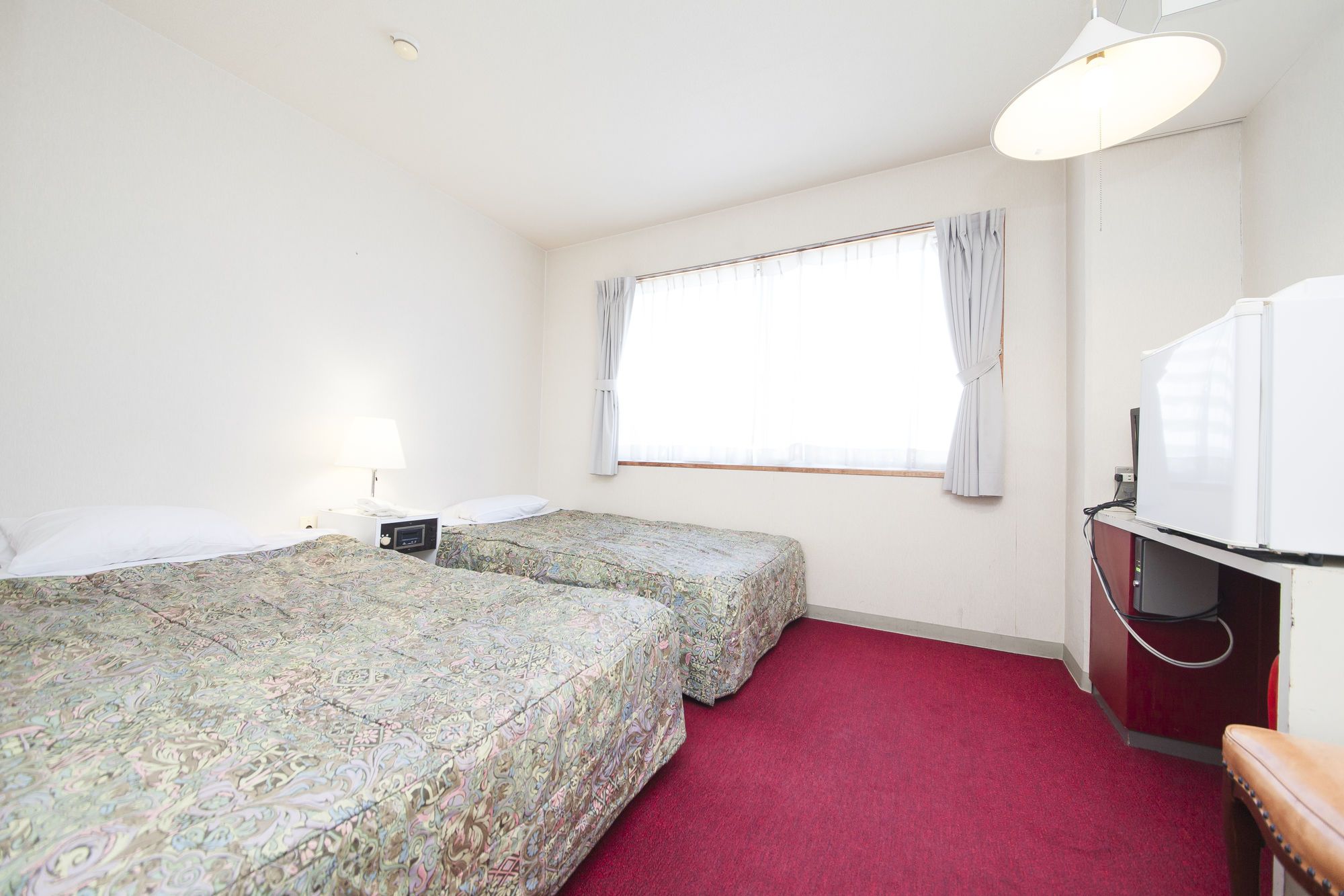 A hotel room with two beds, a desk with a television, a window with curtains, and a red carpet.