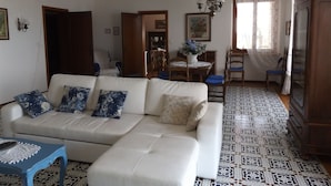 A living room with a white sofa, a blue table, patterned floor tiles, a dining area with a table and chairs, and a chandelier. - Villa Patrizia (Torri del Benaco)