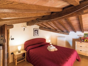 A bedroom with a wooden ceiling, a red bedspread, a bedside table with a lamp, and a wooden dresser with a vase of flowers. - Podere La Cavallara by Interhome (Lago di Bolsena)