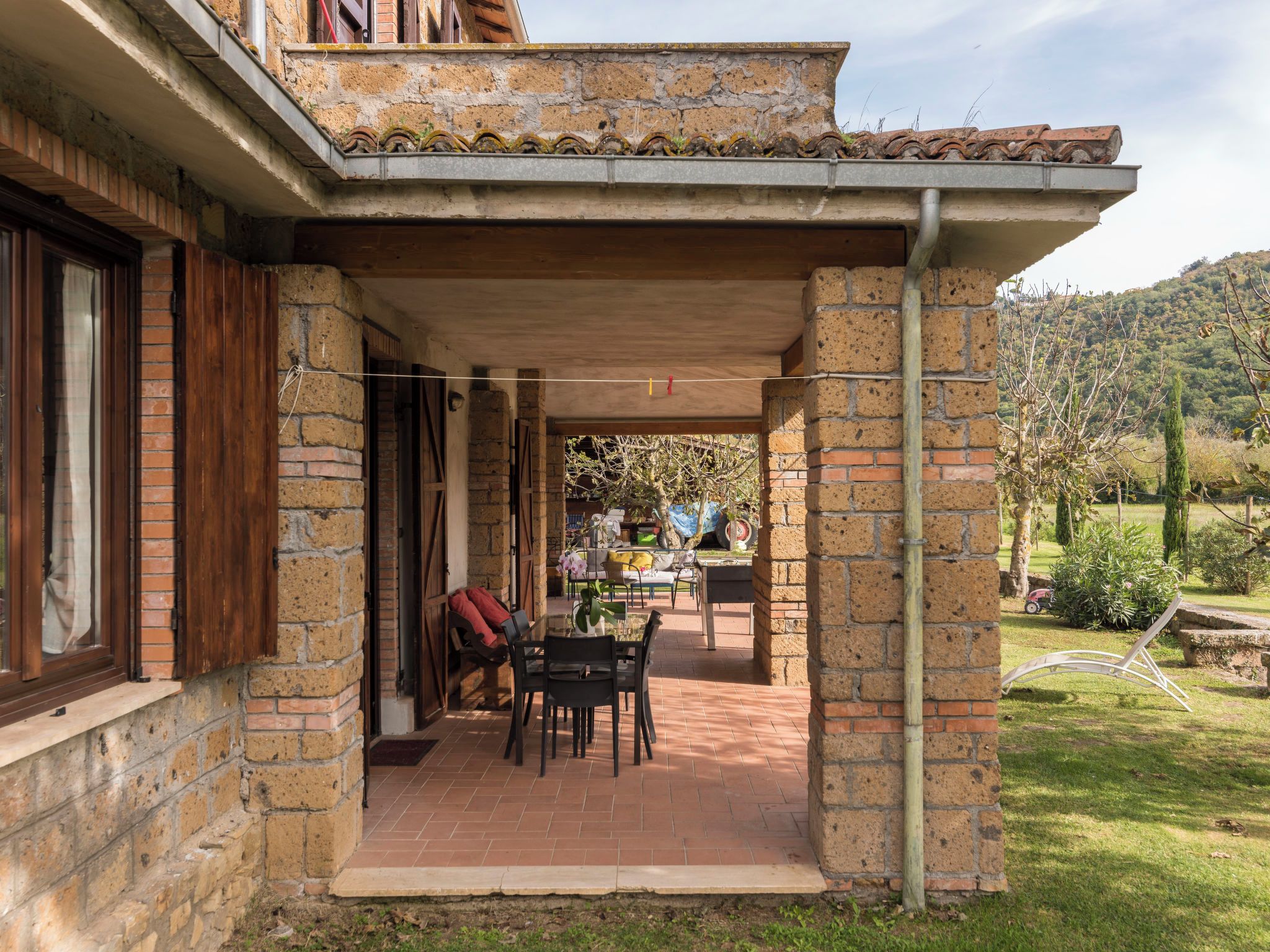 A covered patio area with brick walls, a wooden table and chairs, and a view of a garden and hills in the background.