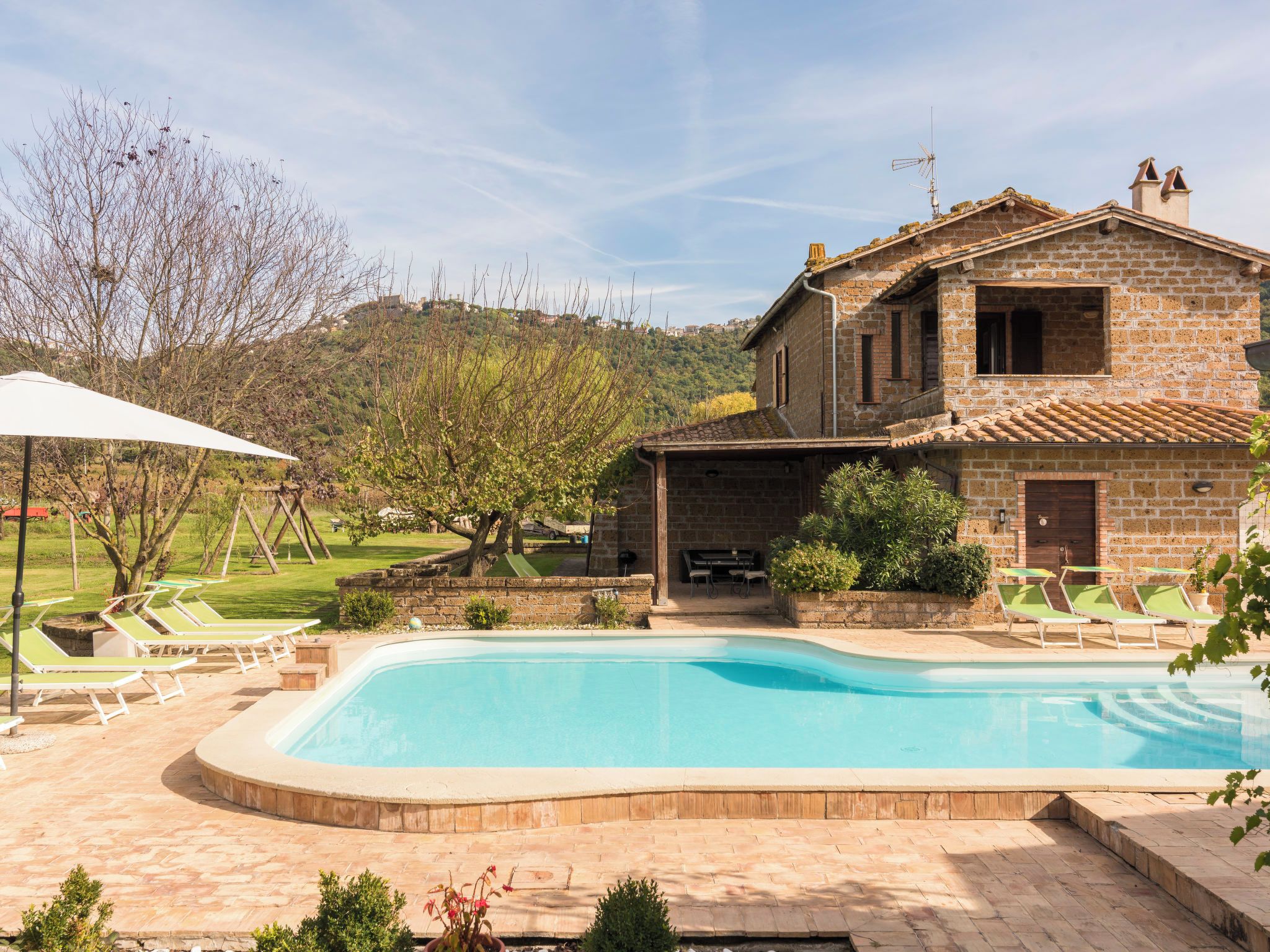A two-story stone house with a swimming pool, outdoor seating, and a view of a hillside.