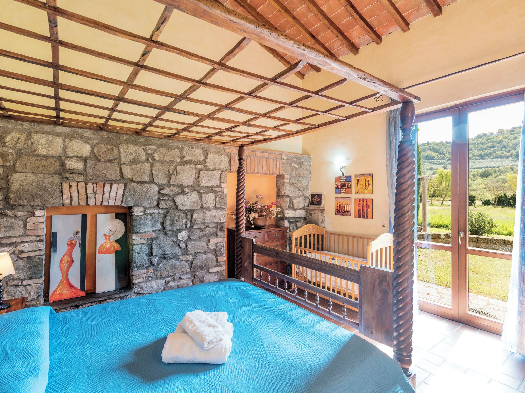 A bedroom with a stone wall, a bed with a blue bedspread, a wooden ceiling, and a balcony with a view.