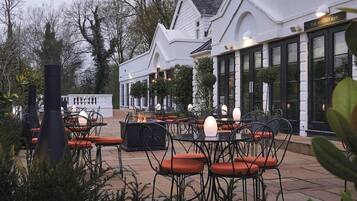 An outdoor seating area with tables and chairs, a fire feature, and a building with a sign reading 'The Monkey Bar'.