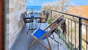 Uma varanda com mesa e cadeiras, uma cadeira dobrável azul e vista para o mar.