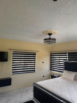 Quarto com cama, lustre e duas janelas com persianas.