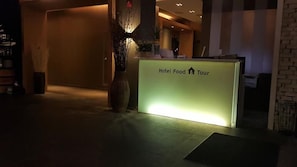 A reception desk with a sign reading 'Hotel Food Tour' in a dimly lit lobby with a vase of dried plants and a carpeted floor. - Hotel Food & Tour (Gwangju)