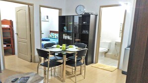 A compact room with a dining table set for two, a black bookshelf, a bed with a colorful bedspread, and a toilet.