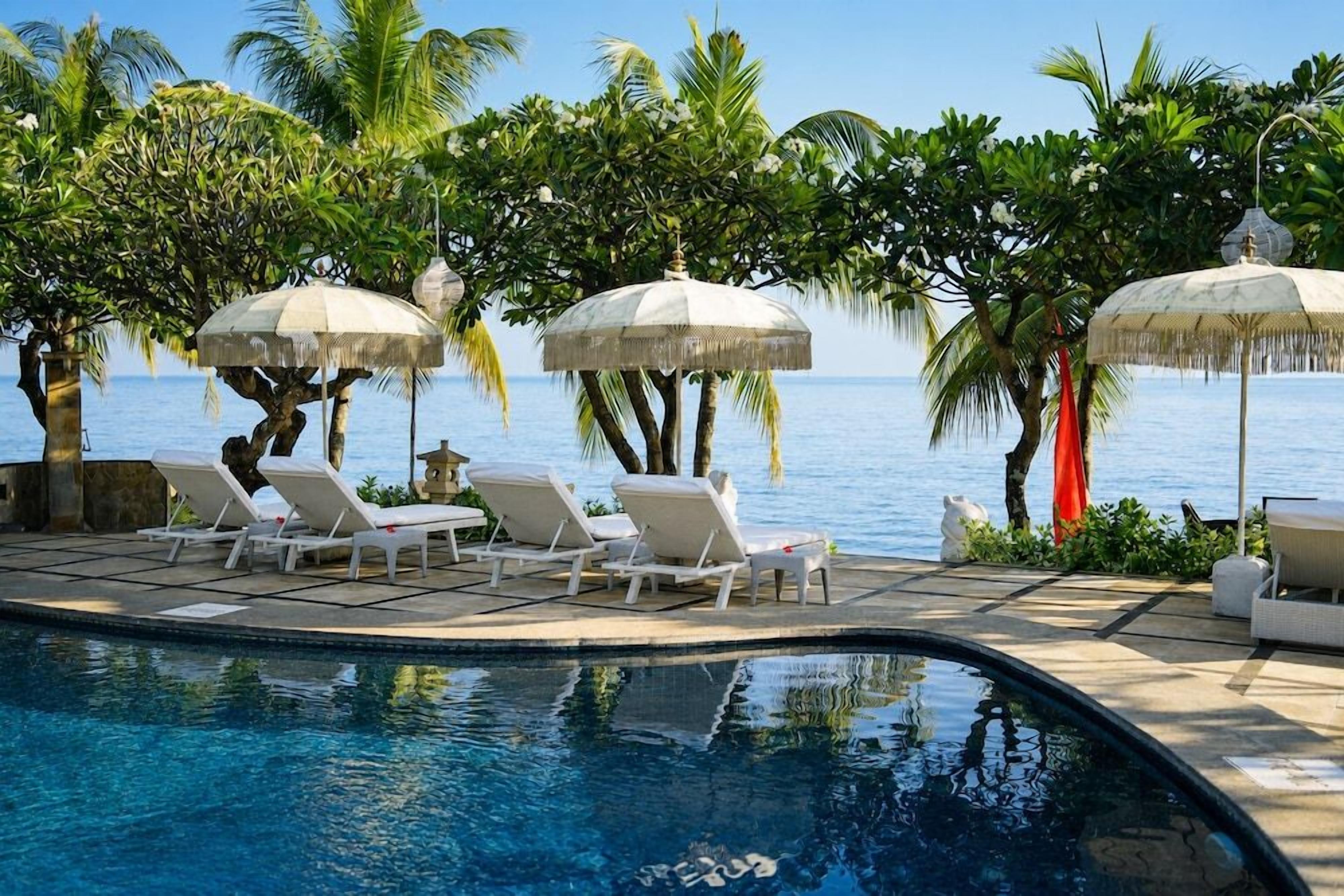 A poolside area with lounge chairs and umbrellas, surrounded by palm trees and a view of the sea.