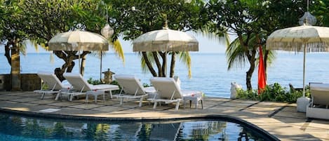 A poolside area with lounge chairs and umbrellas, surrounded by palm trees and a view of the sea.