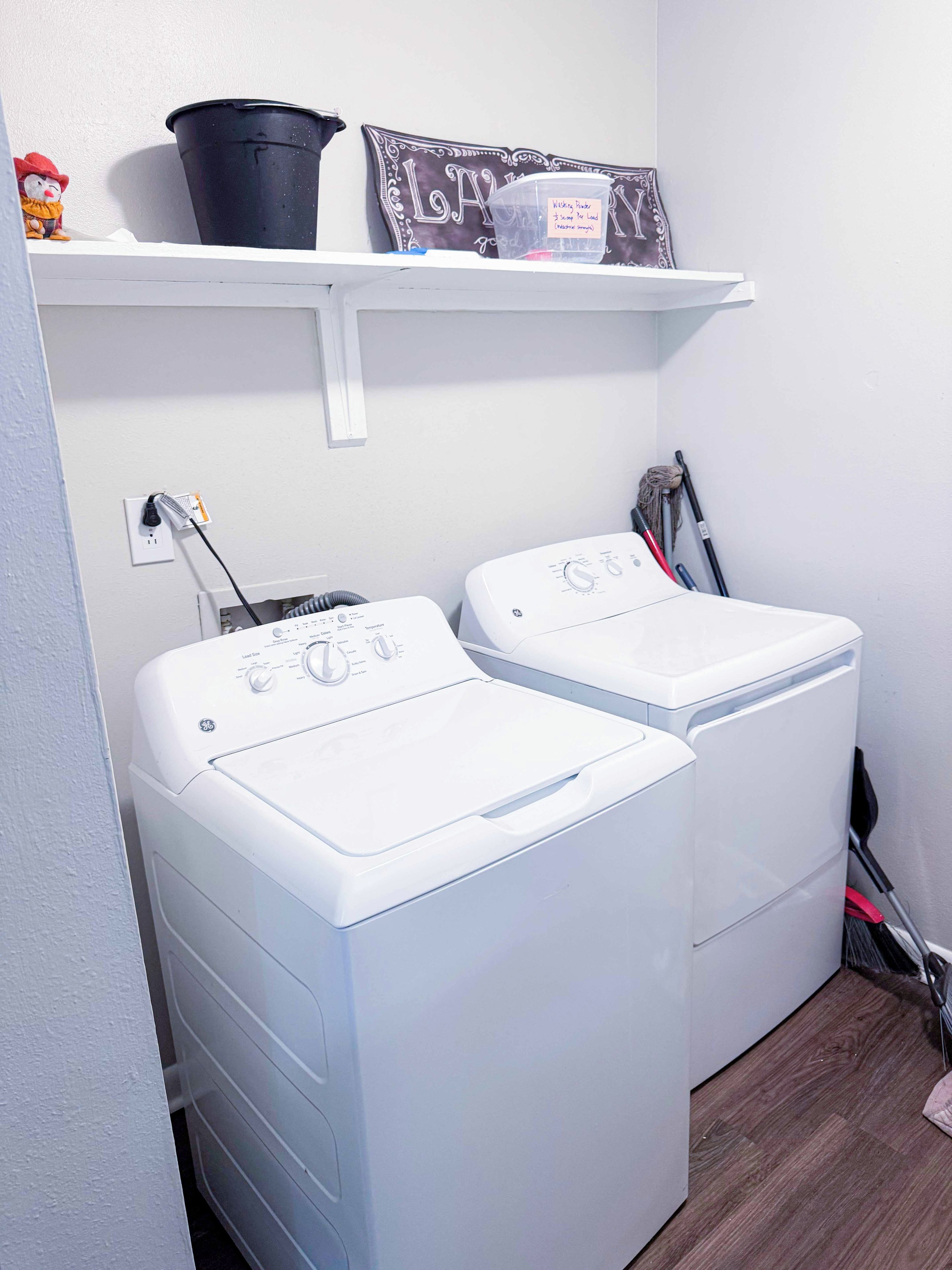 A laundry area with two front-loading washing machines, a shelf with a black bucket and a decorative sign, and a mop leaning against the wall.