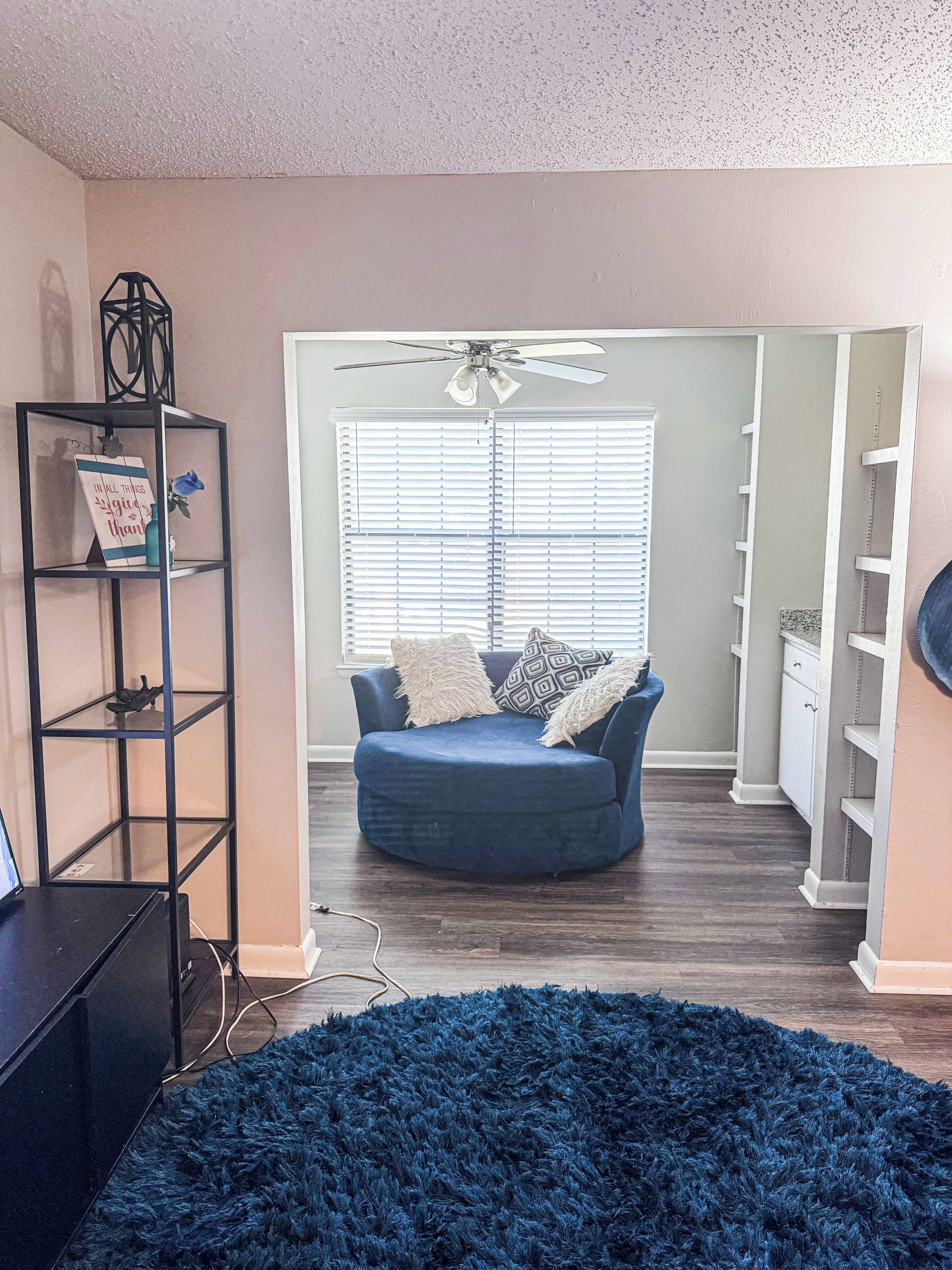 A room with a blue sofa, a ceiling fan, a bookshelf, and a large blue rug.