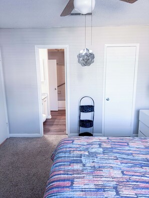 Ein Schlafzimmer mit Bett, Deckenventilator, einer Tür und einem zur Toilette führenden Gang.