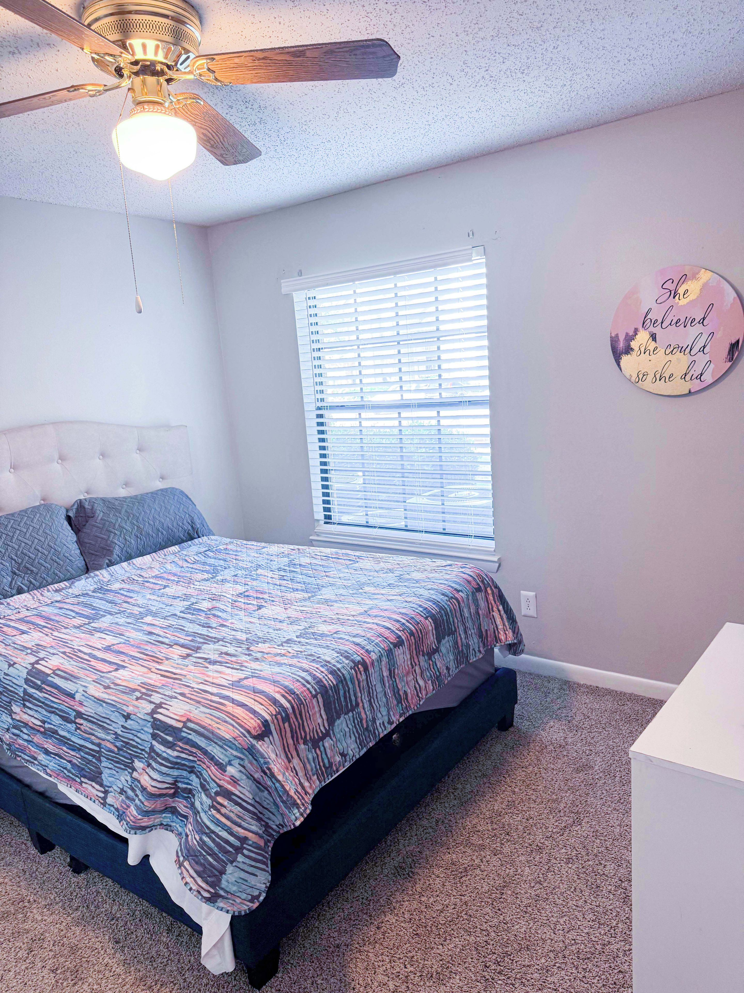 A bedroom with a bed, a ceiling fan, a window with blinds, and a motivational quote on the wall.