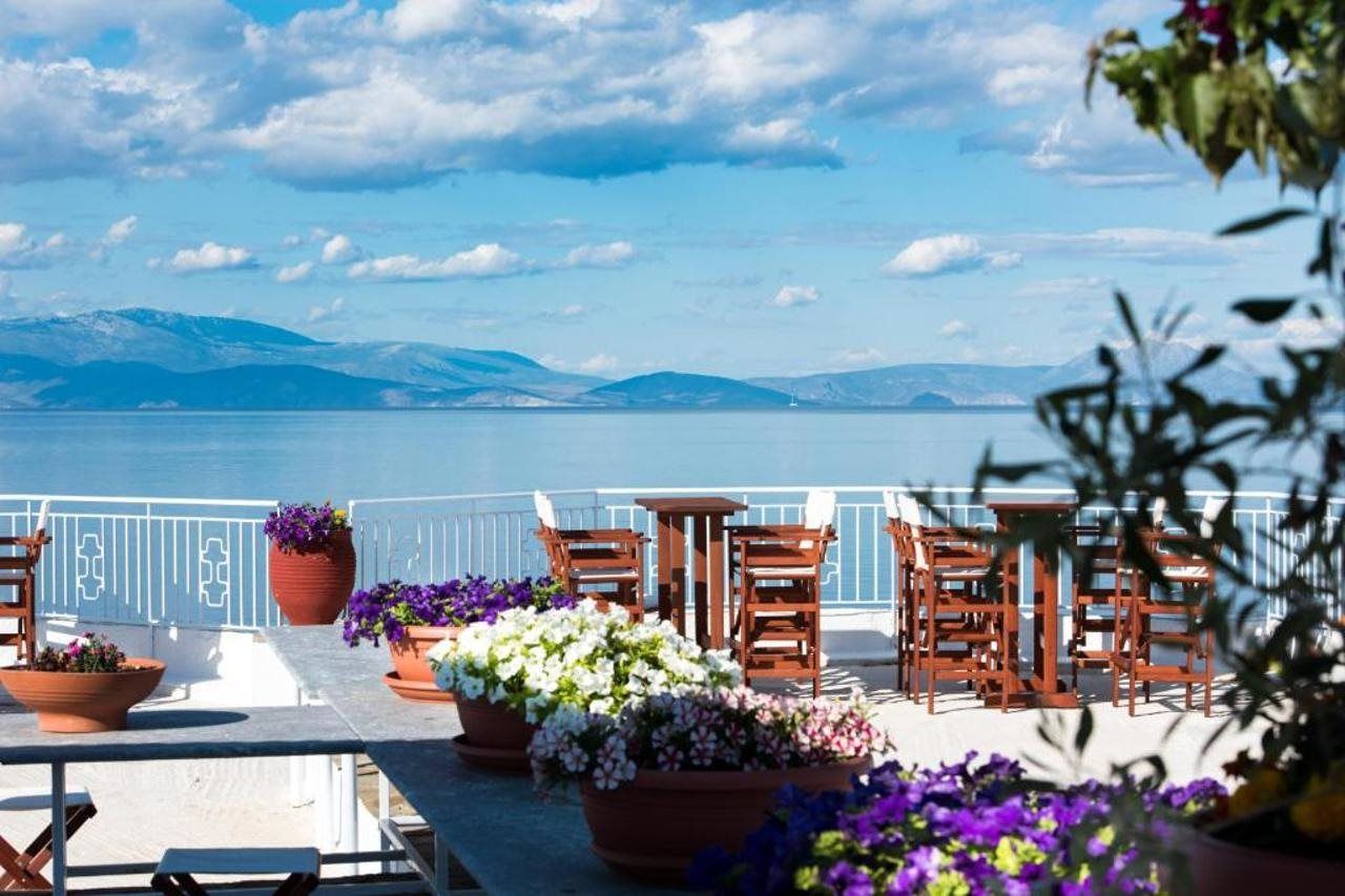 An outdoor terrace with a view of a body of water and mountains, furnished with wooden chairs and tables, and surrounded by potted plants.