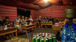 A communal dining area with people seated at tables, a bowl of green cans, and water bottles on the floor. - Martian 2 (Wadi Rum)