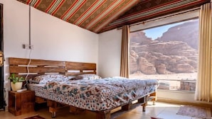 A bedroom with a wooden bed, a potted plant, and a view of a rocky landscape through a large window. - Martian 2 (Wadi Rum)