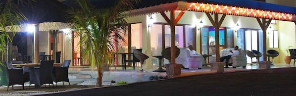 An outdoor seating area with tables and chairs, a covered section with a patterned roof, and a person seated. - Orchid Villas Mauritius (Belle Mare)
