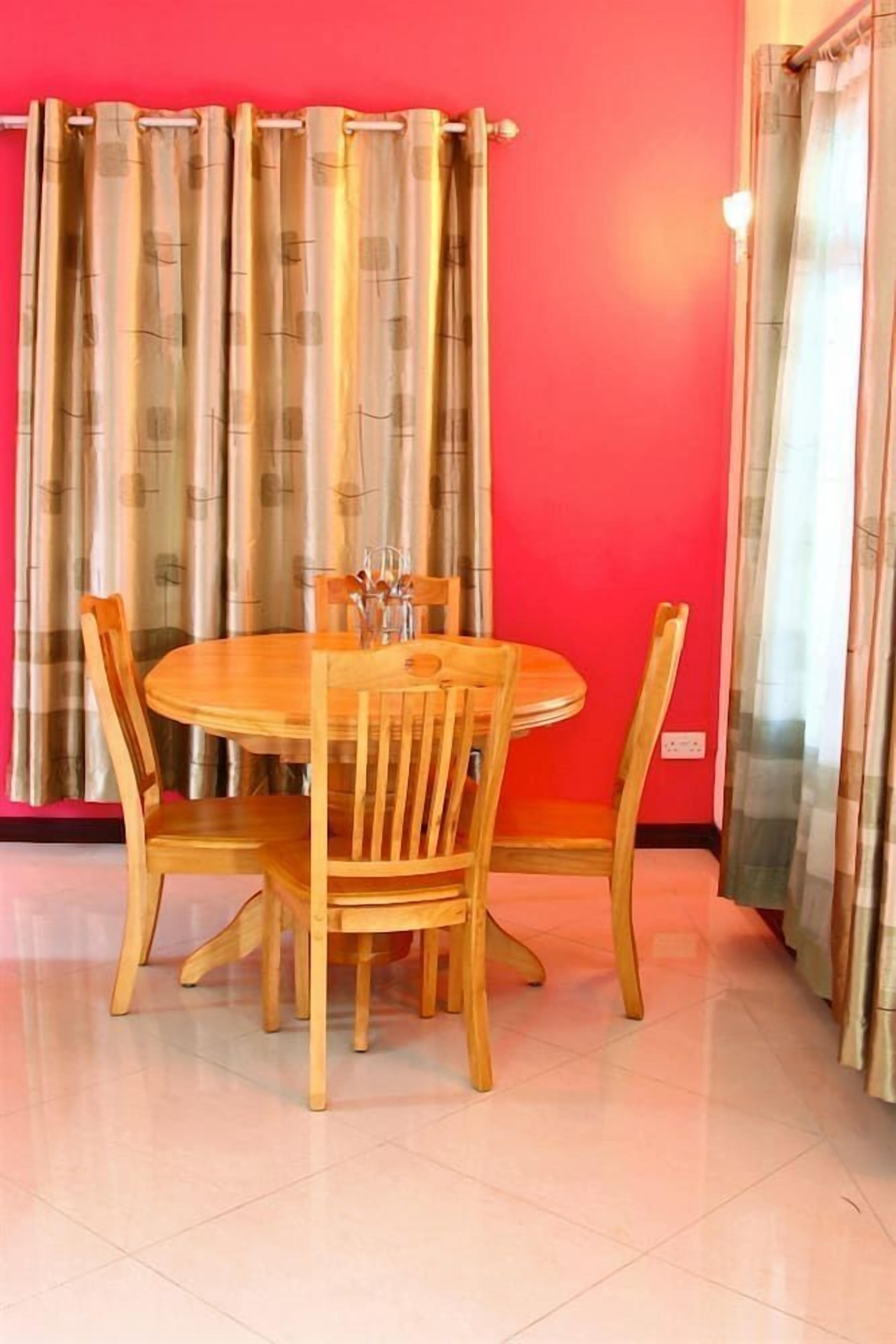 Un comedor con una mesa redonda de madera y sillas, una pared roja y cortinas.