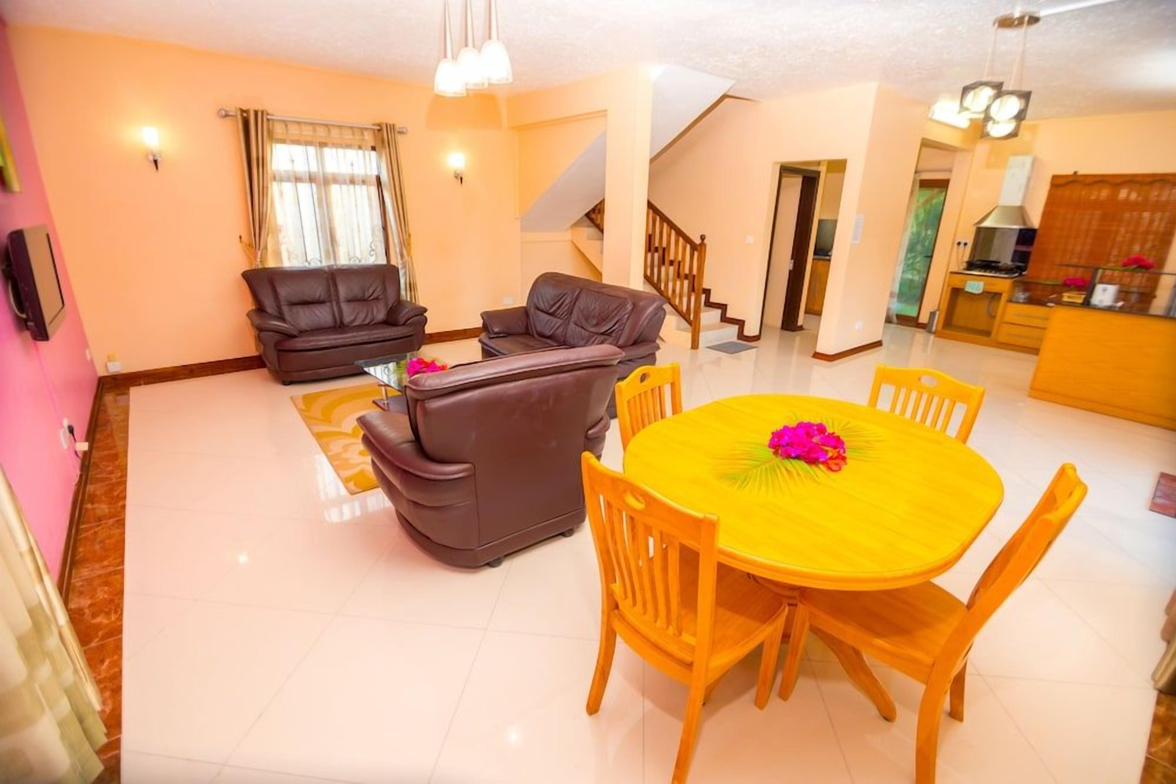 A living room with a sofa and chairs, a dining table with chairs, a staircase, and a kitchen area.