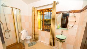 Un baño con cabina de ducha de vidrio, inodoro, lavamanos de vidrio y ventana con cortinas.